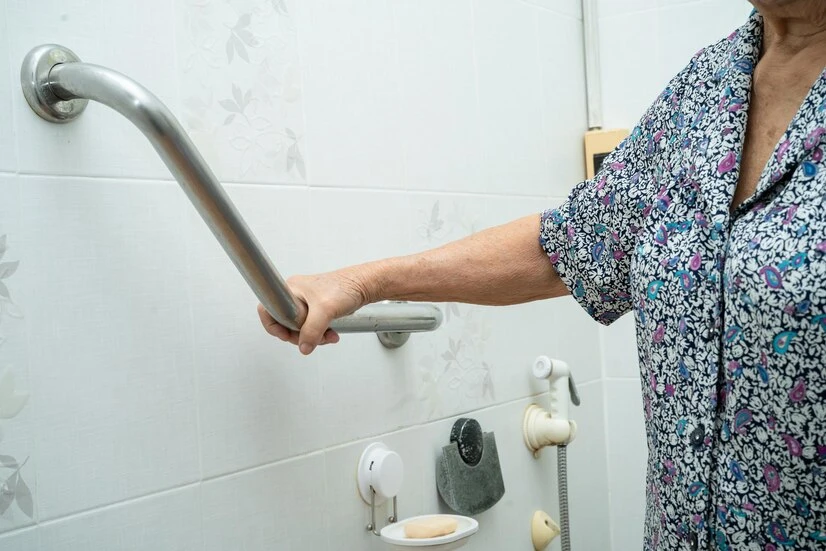 Imagem de uma barra de apoio no banheiro sendo utilizada por uma pessoa idosa. A barra proporciona segurança e estabilidade para o uso do banheiro, essencial na prevenção de quedas.