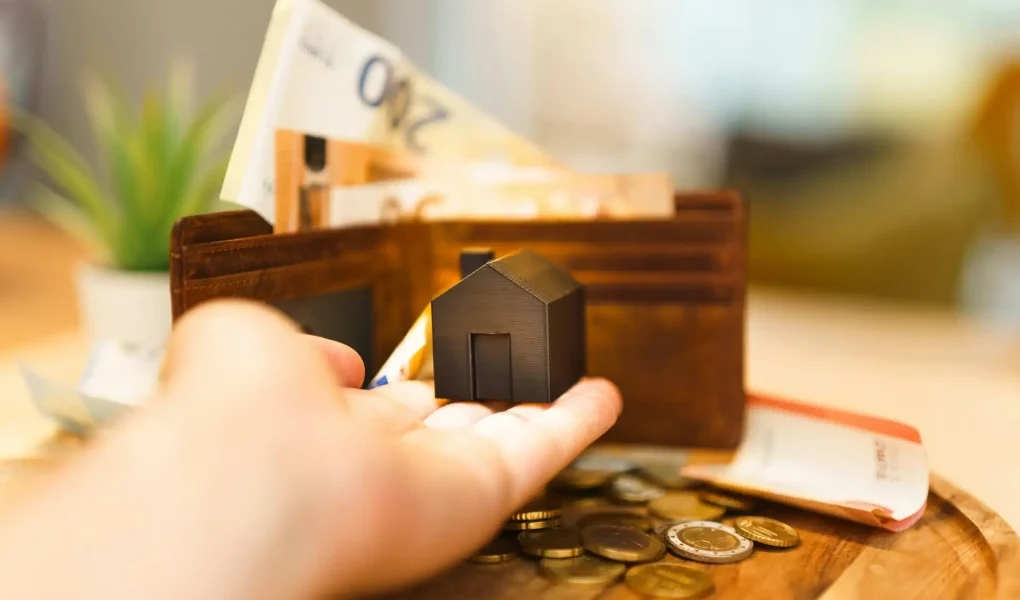 Imagem de uma mão segurando uma pequena casa de modelo, com uma carteira de dinheiro e moedas ao fundo, relacionada a investimentos em fundos imobiliários (FIIs).
