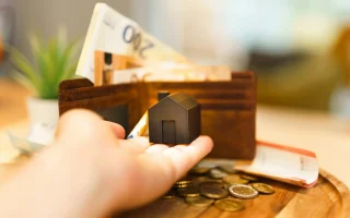 Imagem de uma mão segurando uma pequena casa de modelo, com uma carteira de dinheiro e moedas ao fundo, relacionada a investimentos em fundos imobiliários (FIIs).