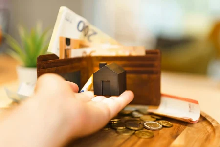Imagem de uma mão segurando uma pequena casa de modelo, com uma carteira de dinheiro e moedas ao fundo, relacionada a investimentos em fundos imobiliários (FIIs).