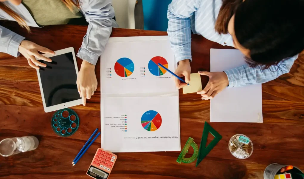 Reunião de profissionais analisando gráficos de tecnologia e orçamento empresarial, com tablet, calculadora e materiais de escritório sobre uma mesa de madeira, ilustrando dicas de como investir em tecnologia sem comprometer o orçamento da empresa.