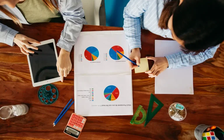 Reunião de profissionais analisando gráficos de tecnologia e orçamento empresarial, com tablet, calculadora e materiais de escritório sobre uma mesa de madeira, ilustrando dicas de como investir em tecnologia sem comprometer o orçamento da empresa.