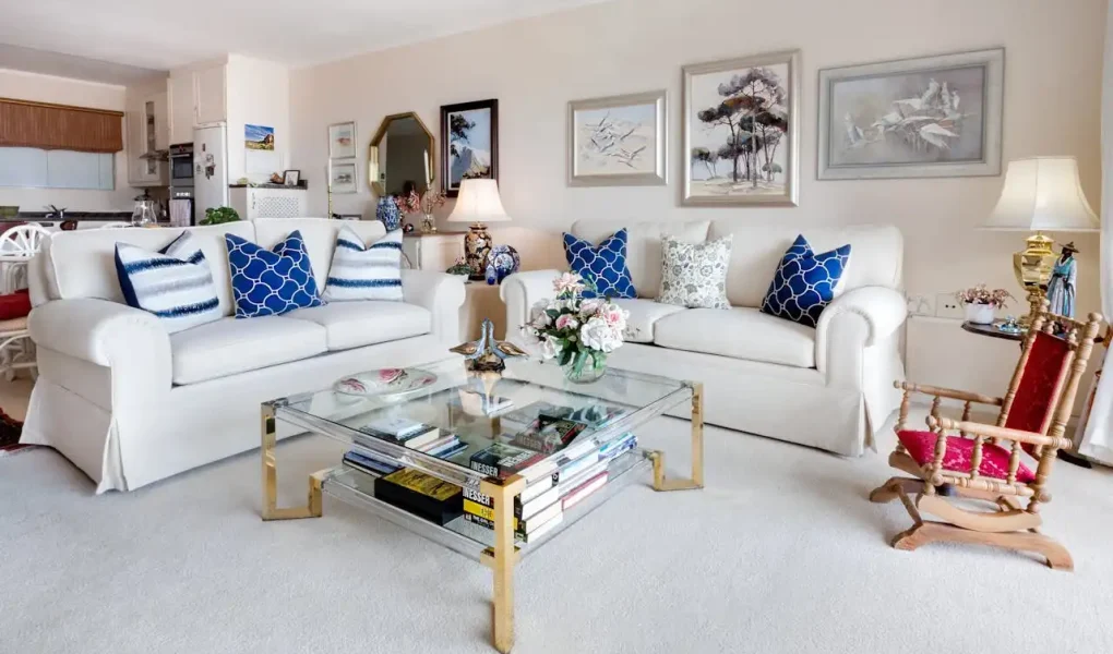 Sala de estar decorada com sofás brancos, almofadas azuis e uma parede com quadros de paisagens, destacando uso de espelhos, luzes e cores para ampliar visualmente o espaço.