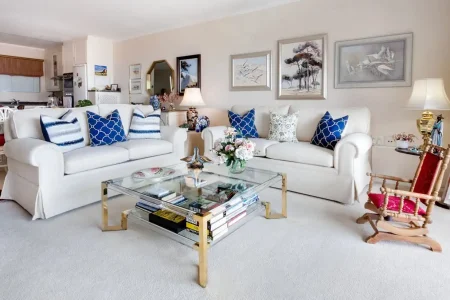 Sala de estar decorada com sofás brancos, almofadas azuis e uma parede com quadros de paisagens, destacando uso de espelhos, luzes e cores para ampliar visualmente o espaço.